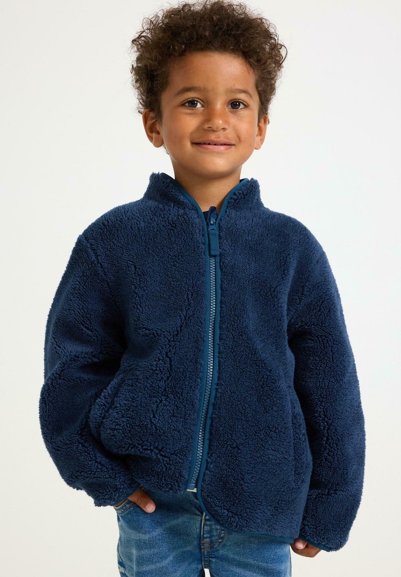Marineblaue Fleecejacke mit Reißverschluss, hohem Kragen und strukturiertem Oberfläche. Kombination mit Denim-Jeans, bietet eine lockere Passform.