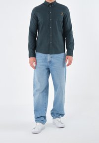 Uomo con camicia button-up verde scuro, jeans azzurri larghi e sneakers bianche, in piedi contro uno sfondo bianco semplice.