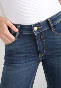 Jeans de mezclilla azul oscuro con un diseño ajustado, que cuentan con un cierre de botón de latón y costuras visibles a lo largo de los bolsillos y las costuras.