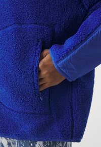 Blå fleecejacka med en strukturerad, fluffig yta. Har en sidoficka och slätt tyg på ärmarna. Närbild av armen.