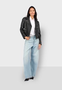 Mujer de pie contra un fondo liso, vestida con una chaqueta de cuero negra, camisa blanca, pantalones vaqueros de pierna ancha azul claro y tacones negros.