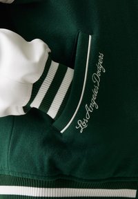 Varsity μπουφάν Los Angeles Dodgers σε σκούρο πράσινο και λευκό. Διαθέτει μάλλινο σώμα, δερμάτινα μανίκια και ριγέ ριμπ μανσέτες. Κεντημένο λογότυπο.