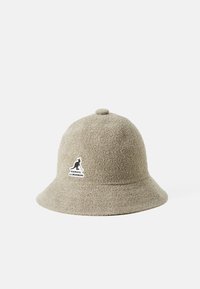 Beige bøttehatt laget av teksturert stoff, med en rund krone og en flat skjerm. Inkluderer et svart logopatch foran.