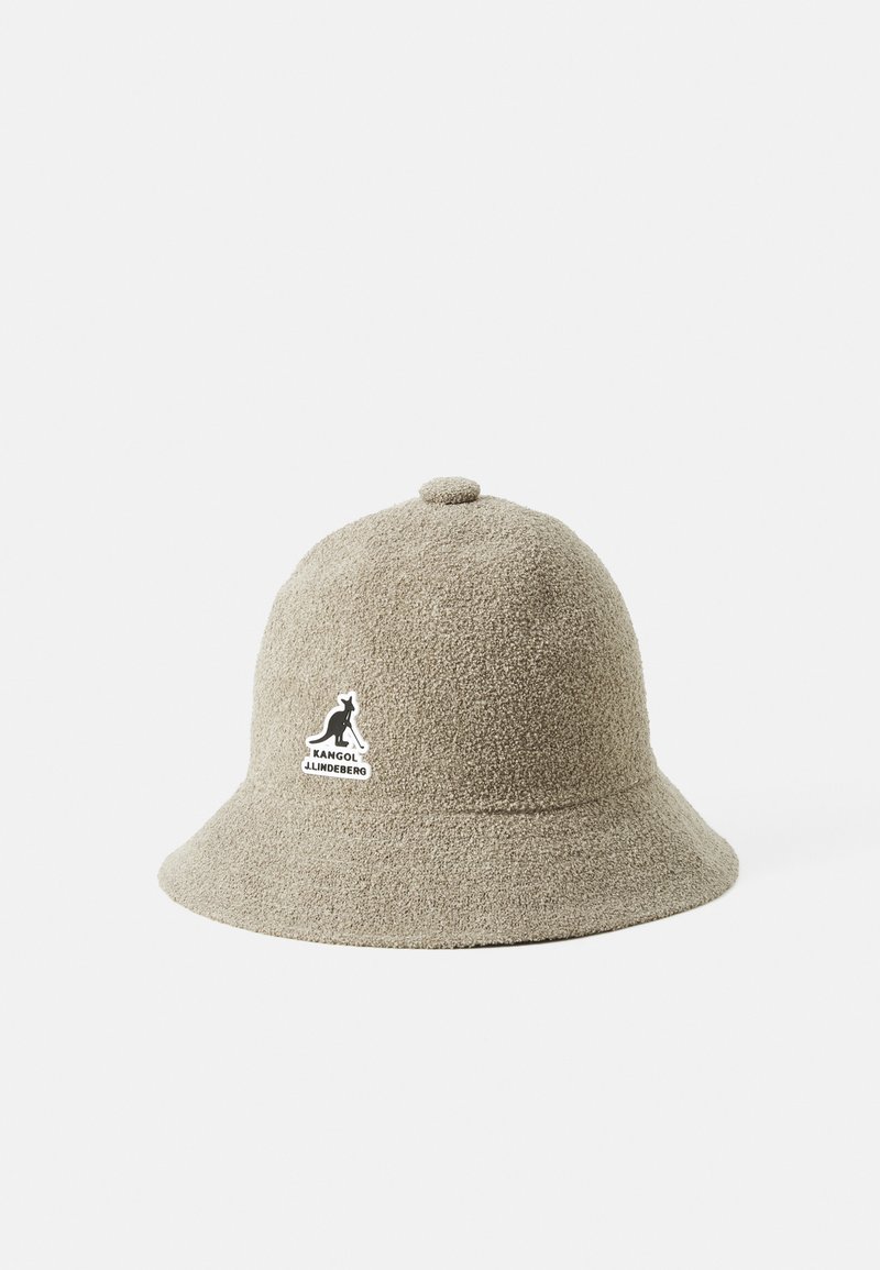 Beige bøttehatt laget av teksturert stoff, med en rund krone og en flat skjerm. Inkluderer et svart logopatch foran.