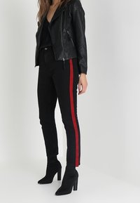 Černá kožená bunda s zipovými detaily, v kombinaci s černými džíny s vysokým pasem, které mají červené boční pruhy, a černými koženými kotníkovými botami na podpatku.