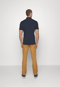 Man som står med ryggen mot kameran, iförd en navyblå kortärmad polo, beige byxor och svarta sneakers mot en enkel vit bakgrund.