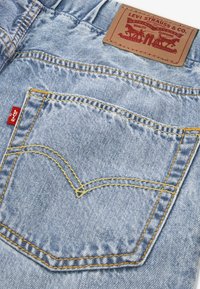 Hellblaue Jeans aus Denim mit einer auffälligen Gesäßtasche, gelben Nähten und einem roten Logoetikett. Der Bund ist mit einem braunen Lederpatch versehen.