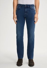Jean en denim bleu foncé avec une coupe droite, passants de ceinture, poches avant et une texture discrète. Porté avec des bottes marron foncé.