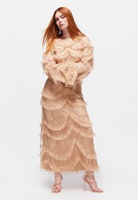 Beige mekko, jossa on kerroksellisia röyhelöitä, läpinäkyvää kangasta ja kukkakuoseja. Ominaisuuksiin kuuluvat kellotakkiset hihat ja pitkä siluetti. Yhteensopivat remmikengät.