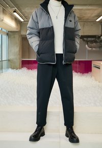 Vadderad jacka i grått och svart färgblockering, med hög krage och dragkedjestängning. Kombinerad med svarta byxor och klumpiga svarta stövlar.