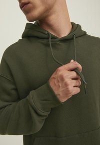 Olivgrüner Hoodie mit Kordelzug, gerippten Bündchen und Kängurutasche. Der Stoff wirkt weich und hat eine glatte Textur.