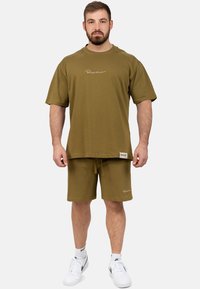 T-shirt in cotone verde oliva con maniche corte e logo ricamato, abbinato a pantaloni corti coordinati con coulisse regolabile. Sneakers bianche.