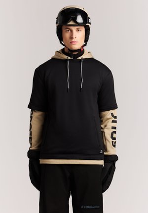Jonge man die een zwarte hoodie met korte mouwen draagt over een beige shirt met lange mouwen, zwarte handschoenen, zwarte broek en een beige skihelm met bril.