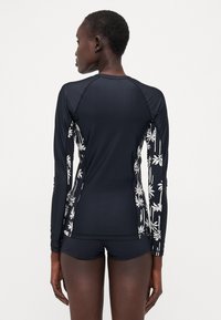 Svart langermet badebluse med hvite palmebladmønstre på sidene. Glatt, elastisk stoff og en tettsittende design. Kombinert med svarte shorts.