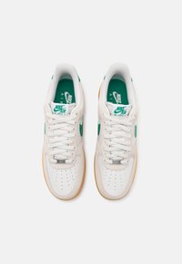 Ténis Nike Air Force 1 brancos com detalhes em verde, apresentando um cabedal em pele suave, caixa de pé perfurada e uma sola em borracha de côr âmbar.