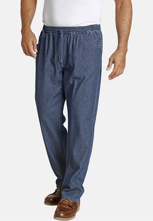 Mann, der blaue Jeans mit elastischem Bund und Kordelzug sowie braune Freizeitschuhe trägt, vor weißem Hintergrund stehend.