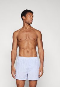 Blå og hvite stripete boksershorts med elastisk midjebånd, lett stoff og knapp detalj foran. Modellen poserer uten skjorte.