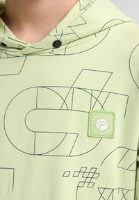 Sudadera verde claro con patrones de líneas geométricas negras. Incluye un parche cuadrado con un logo y botones a presión metálicos en el cuello.