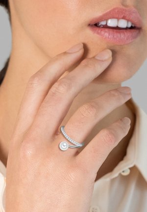 Anillo de plata con una perla y banda contrastante con piedras claras, posado sobre una mano que toca la barbilla. Acabado metálico suave.