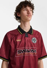 Jonge man met krullend haar die een bordeaux sportshirt draagt met zwarte en gele bies, met de merken "SWARM Internationals" en "hummel sportswear."