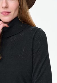 Vrouw met lang lichtbruin haar, gekleed in een donkere coltrui met een subtiel breipatroon en een bruine hoed, tegen een witte achtergrond.