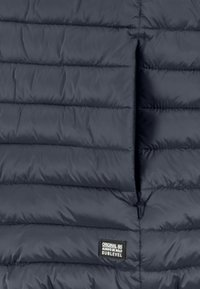 Nahaufnahme einer dunkelblauen steppjacke mit horizontalen Nähten, die eine Taschenerweiterung und ein kleines Stoffmarkenschild zeigt.