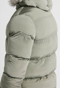 Giacca imbottita verde chiaro con sezioni trapuntate orizzontali, cappuccio foderato in morbida pelliccia e texture liscia e riflettente.