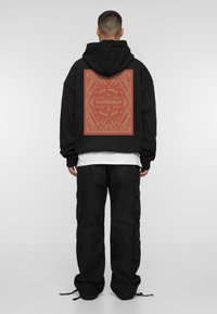Black hoodie with a large red graphic on the back featuring intricate patterns and text; paired with loose-fitting black cargo pants.