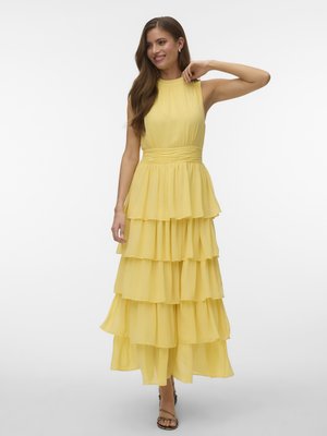 Femeie într-o rochie maxi galbenă fără mâneci, cu volane și tocuri negre cu barete, zâmbitoare și atingându-și părul pe un fundal alb simplu.