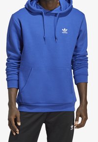 Sininen huppari, jossa on etutasku ja valkoinen logo. Valmistettu pehmeästä kankaasta, jossa on ribbiohut hihansuissa ja vyötäröllä, sileä pinta.