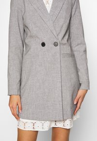 Szary, podwójnie zapięty żakiet z czarnymi guzikami, gładka faktura, klapy typu notch oraz kieszenie boczne, noszony na białej koronkowej sukience.