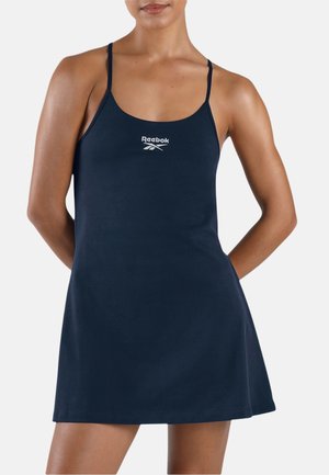 Vrouw die een mouwloze marineblauwe Reebok-jurk draagt met dunne bandjes en een logo in het midden van de borst, tegen een effen achtergrond.