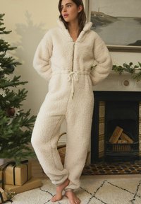 Cremefarbene Fleece-Einteiler mit Reißverschluss vorne, hohem Kragen und Kordelzug in der Taille. Verfügt über ein strukturiertes Material und eine lockere Passform, ideal für Wärme.