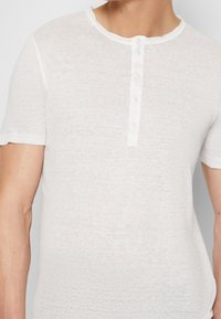 Bílé krátké tričko henley s krátkým rukávem vyrobené z lehkého materiálu, s kulatým výstřihem a čtyřmi předními knoflíky, s mírně texturovaným povrchem.