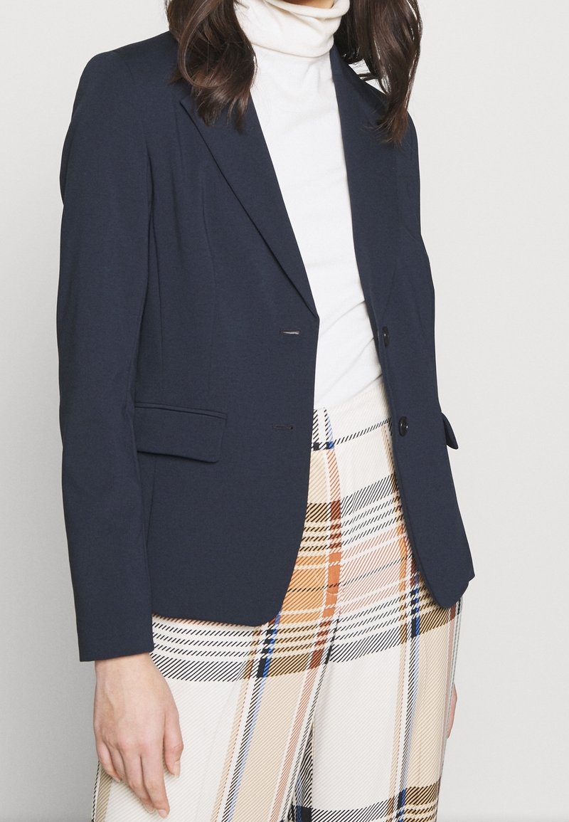Femme portant un blazer bleu marine sur un pull à col roulé blanc et un pantalon taille haute à motif à carreaux beige, orange, bleu et noir.