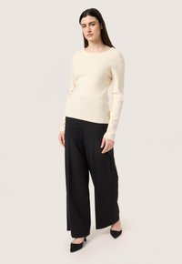 Cremiger Strickpullover mit langen Ärmeln, kombiniert mit weit geschnittenen schwarzen Hosen und schwarzen spitzen Schuhen. Strukturiertes Material und taillierter Schnitt.