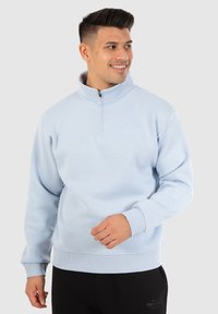 Pull en bleu clair en tissu doux avec un col zippé mi-haut, des poignets côtelés et une coupe légèrement cintrée. Branding minimal sur la poitrine.