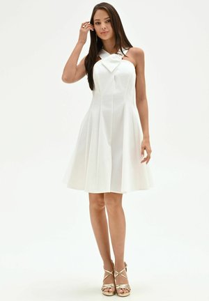 Femme debout portant une robe blanche sans manches avec un détail nœud et des sandales dorées à talons hauts sur fond blanc uni.