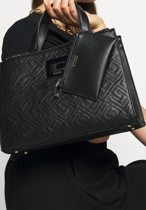 Sac à main en cuir matelassé noir avec motif géométrique et pochette assortie zippée tenue par une personne vêtue de noir et portant un collier en chaîne dorée.
