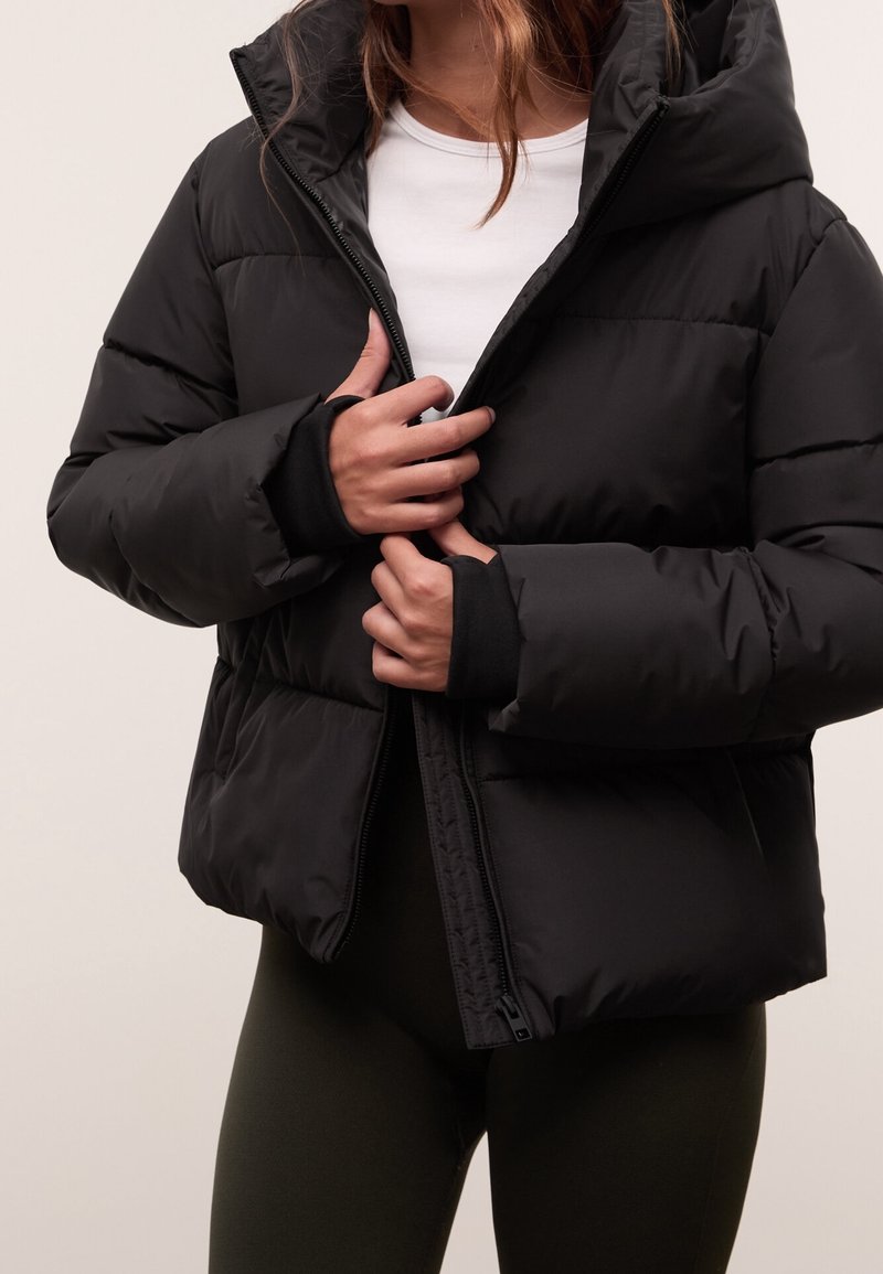 Personne portant une doudoune noire sur une chemise blanche, en train de zipper la veste, avec des leggings foncés visibles de la taille vers le bas.