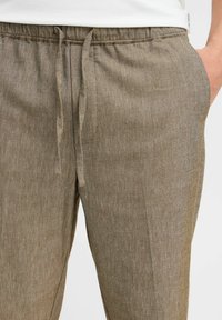 Brown textured drawstring pants with elastic waistband, side pockets, worn with a white shirt visible at the waist.