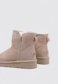 Botines beige de ante con forro de borrego, suela plana y lengüeta en la parte trasera. Presentan una marca sutil en el talón.