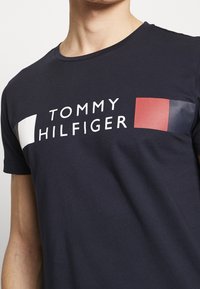 Man wearing navy blue T-shirt with white text "Tommy Hilfiger" and rectangular white, red, and navy color blocks on the chest.