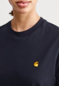Mørkeblå bomulds T-shirt med rund halsudskæring og et lille gult logo på venstre brystområde. Glat tekstur, korte ærmer.