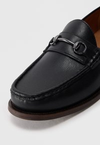 Mocasines de cuero negro con punta redonda, herrajes decorativos en plata y sutiles detalles de costura en la parte superior y la suela.