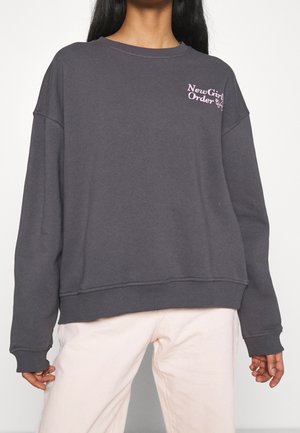Personne portant un sweat-shirt gris foncé avec le texte "New Girl Order" en rose sur la poitrine gauche et un pantalon clair.