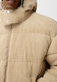 Beige corduroy puffer jacket featuring a hood, vertical ribbed texture, snap closures, and a front zipper. The material appears soft and padded.