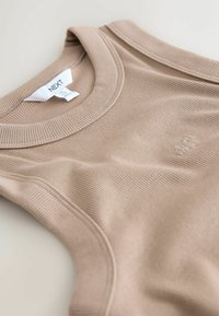 Bézs bordázott trikó kerek nyakkivágással, finom márkajeléssel és megerősített varrásokkal. Puha tapintású, testhez simuló kialakítás.