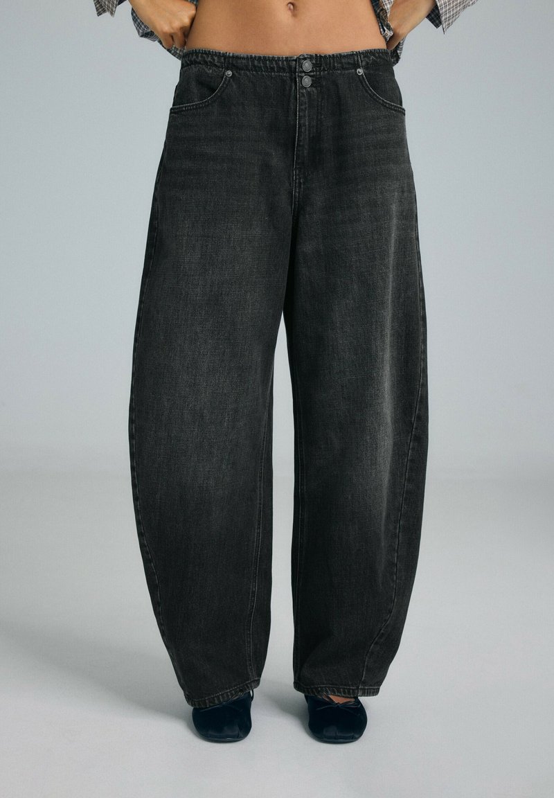 Personne portant un pantalon en jean gris foncé ample, taille haute avec fermeture à boutons, associé à des chaussures plates noires.