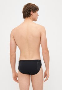 Slip da nuoto neri con una texture liscia e cuciture laterali a contrasto, indossati da una persona vista da dietro, che mette in mostra un design minimalista.
